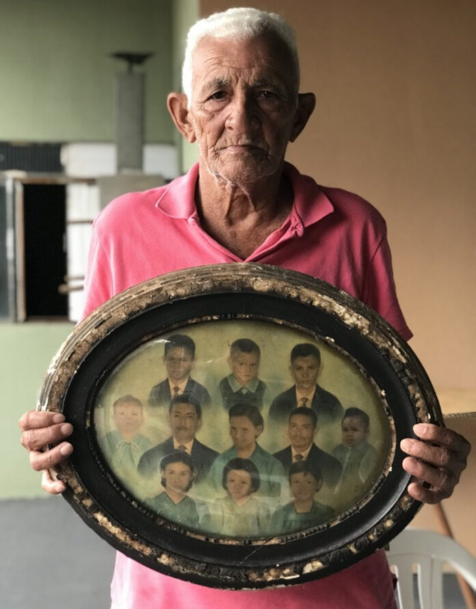 GUARDIÃO DA MEMÓRIA 
– Plínio, em 2023, segura o quadro com fotos de seus pais e irmãos; 
ele aparece na terceira posição da fila superior, 
da esquerda para a direita. 
(Foto: Júlio Olivar)
