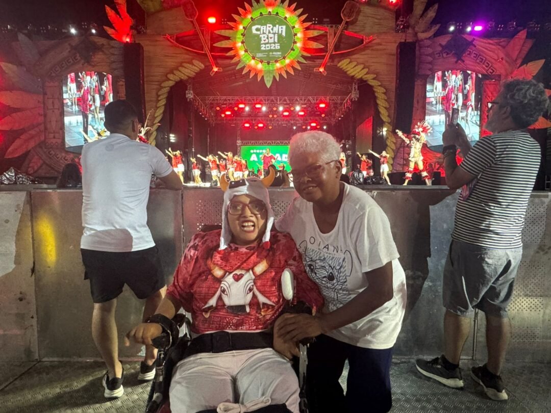Imagem colorida mostra mãe e filha cadeirante pousando para foto. Atrás delas o palco do Carnaboi durante apresentação do Garando